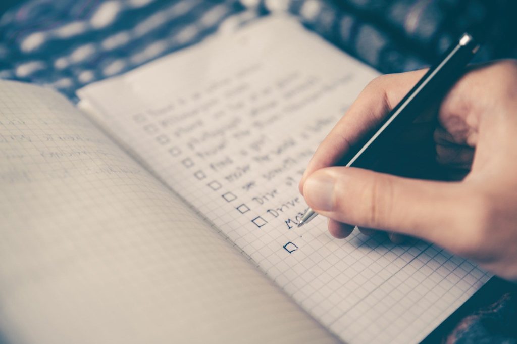 A persons hand holding a pen and writing a list with check boxes on grid paper